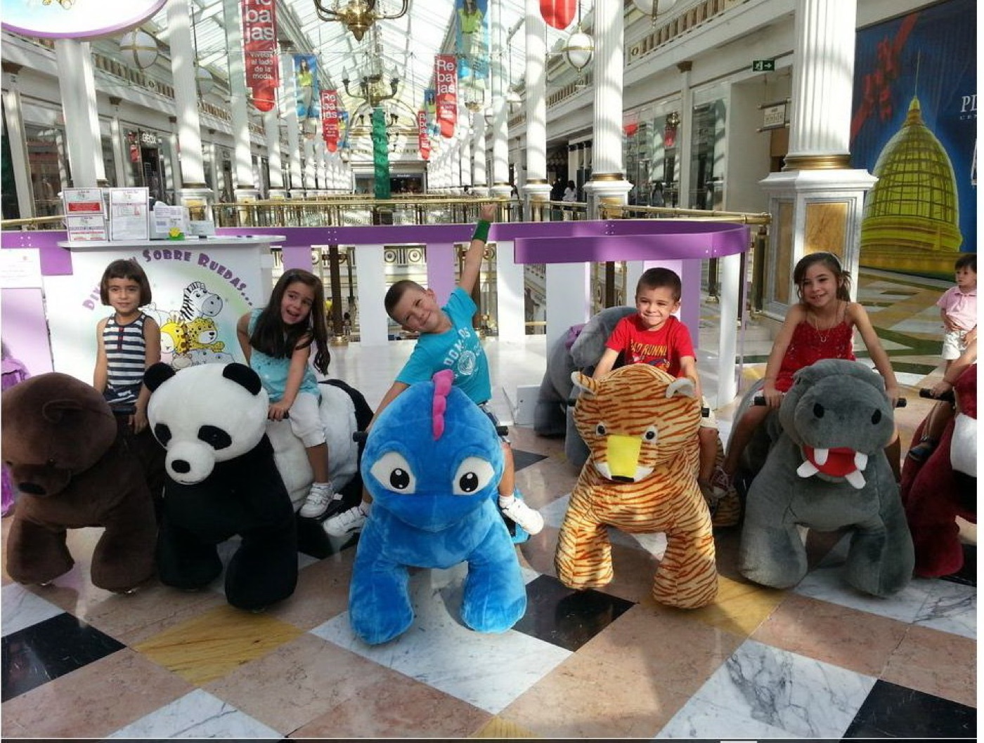 Group of children smiling on motorized plush ride-on animals including a bear, panda, blue dinosaur, and tiger in a bright indoor plaza