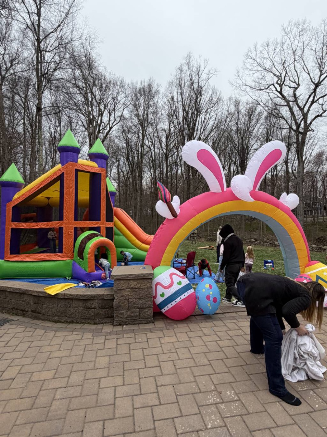 Outdoor party with Easter-themed inflatables: colorful bounce house, large pink arch with bunny ears, oversized Easter eggs, kids and adults on a patio and lawn
